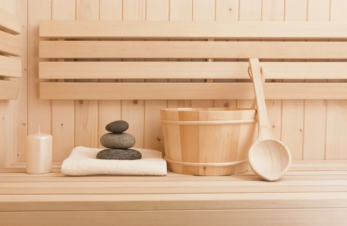 A sauna bench set with a candle, stacked stones, a folded towel, and a wooden bucket and ladle